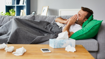 Man, sick at home on his sofa using a tissue to blow his nose while wrapped in a blanket