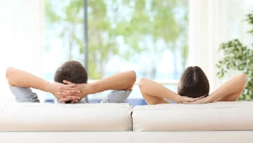 Couple with arms folded behind their heads sitting on a couch looking out a big window