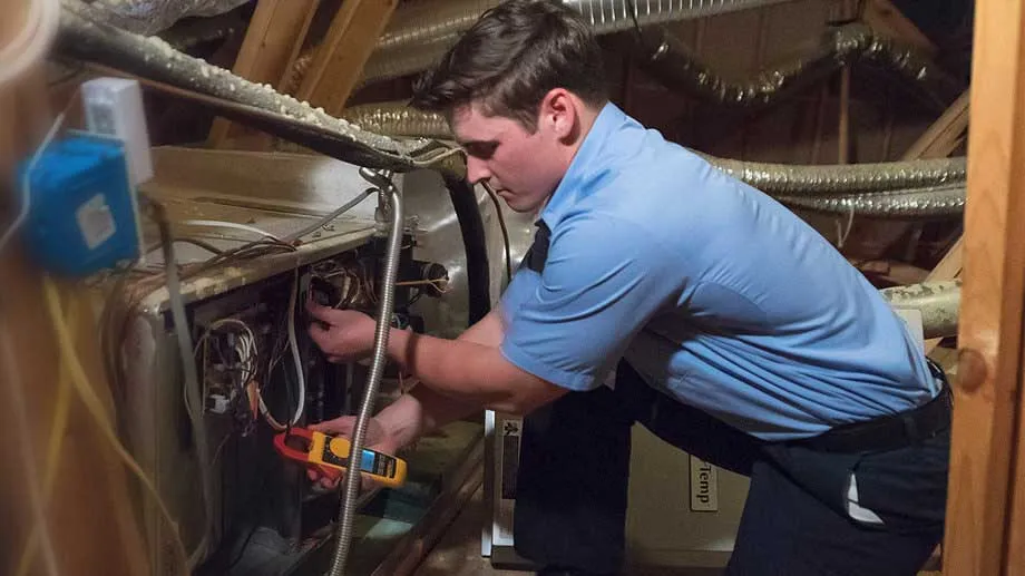Male Aire Serv technician in blue collared shirt holding digital manifold gauge beside exposed furnace.