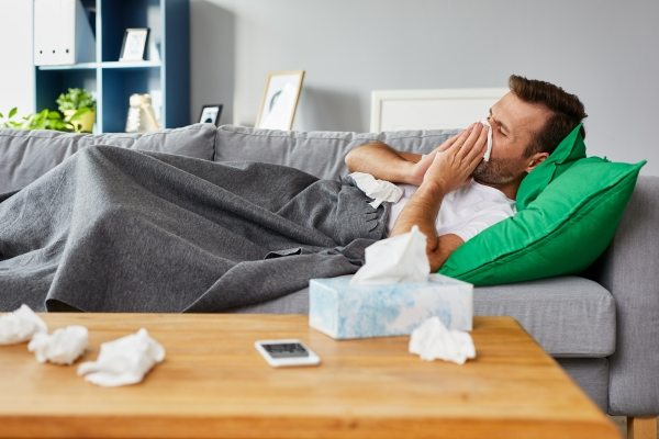Man, sick at home on his sofa using a tissue to blow his nose while wrapped in a blanket
