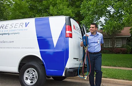 Male Aire Serv technician holding A/C manifold gauge beside parked branded van.
