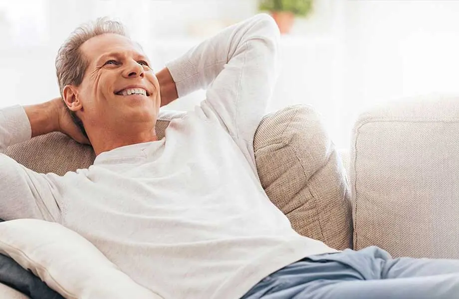 Middle-aged Caucasian man in white sweatshirt smiling and reclining on couch in bright interior.