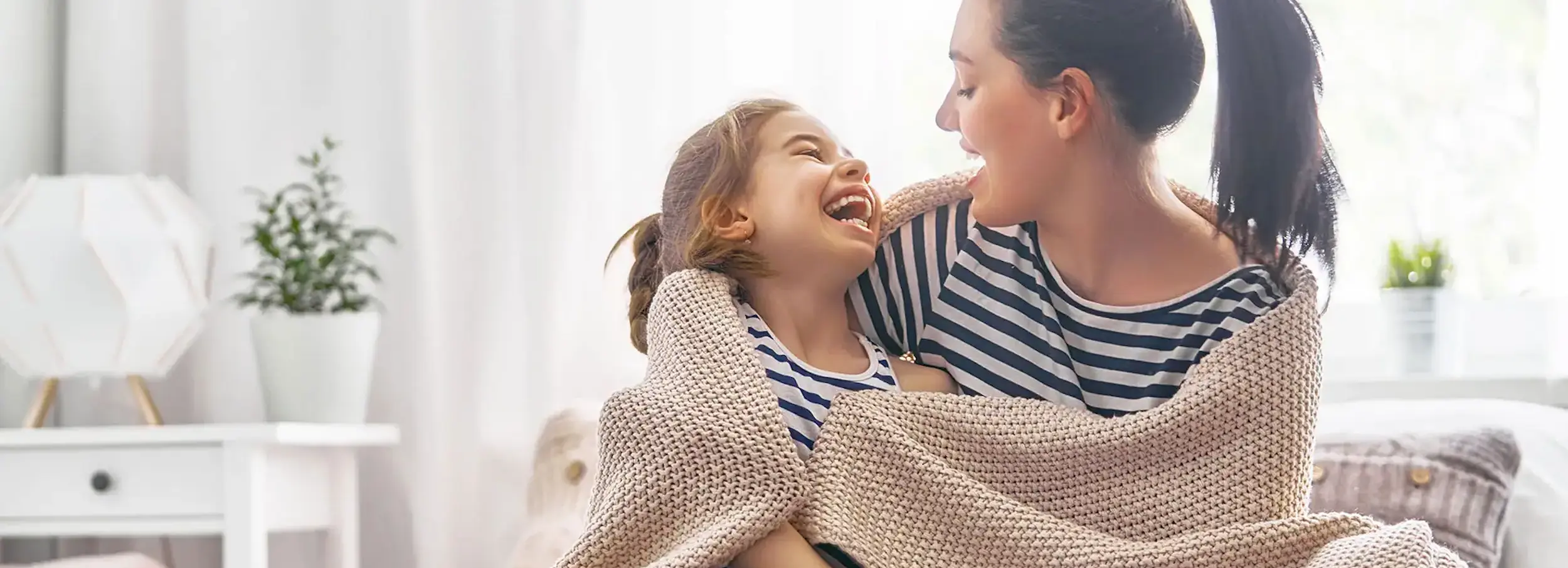 Mother and daughter wrapped in taupe blanket together smiling and laughing in brightly lit living room.